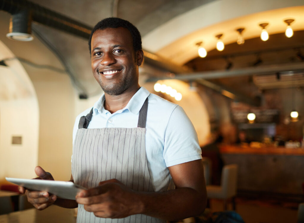 young african american waiter in an apron holding an ipad as he works in a restaurant