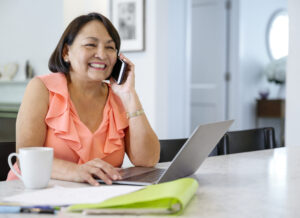 senior woman talking on the phone and using laptop in the kitchen while drinking her coffee and getting 24/7 support